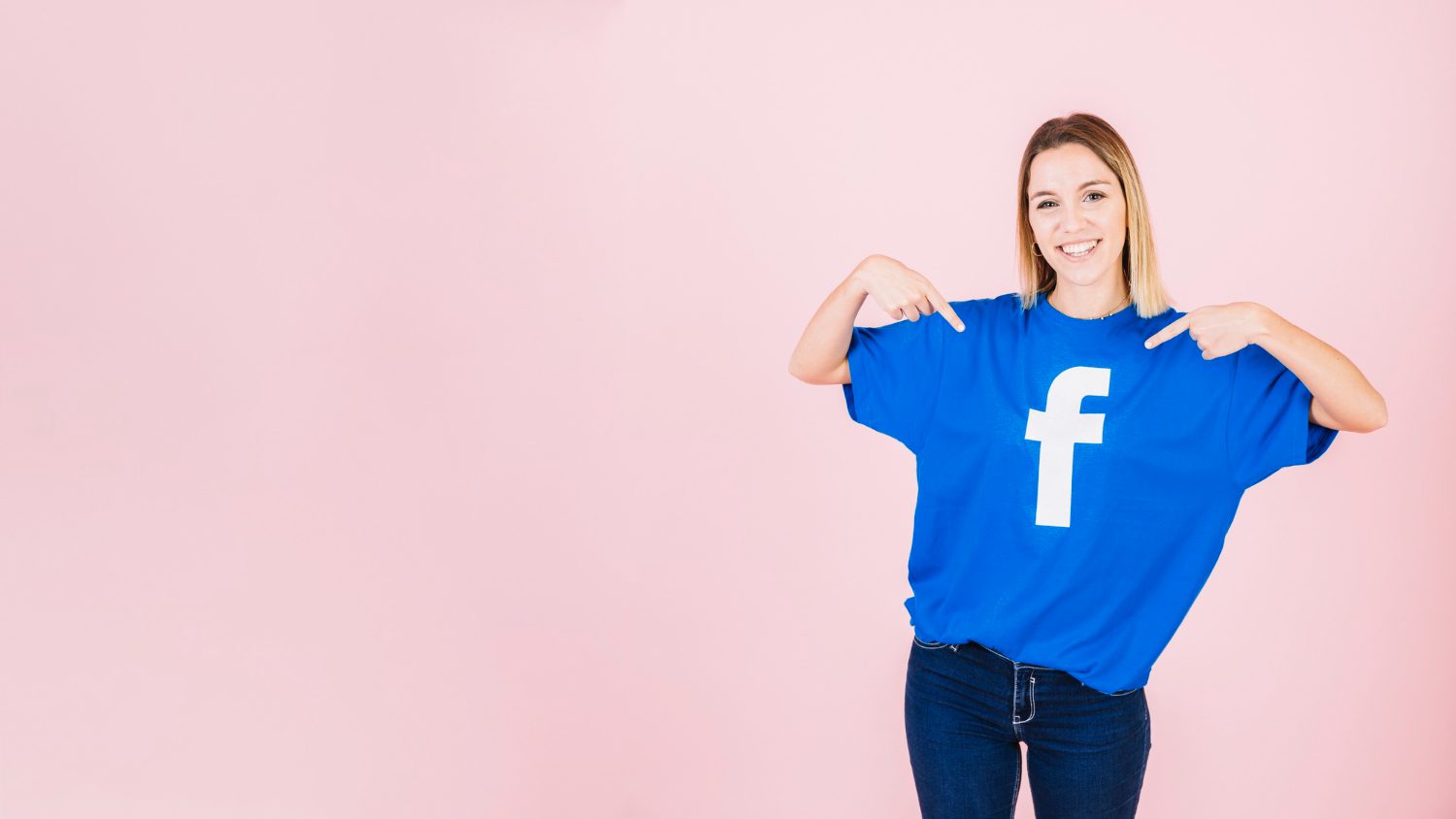 A smiling woman in a blue t - shirt with a white Facebook logo points at it while standing against a soft pink background.