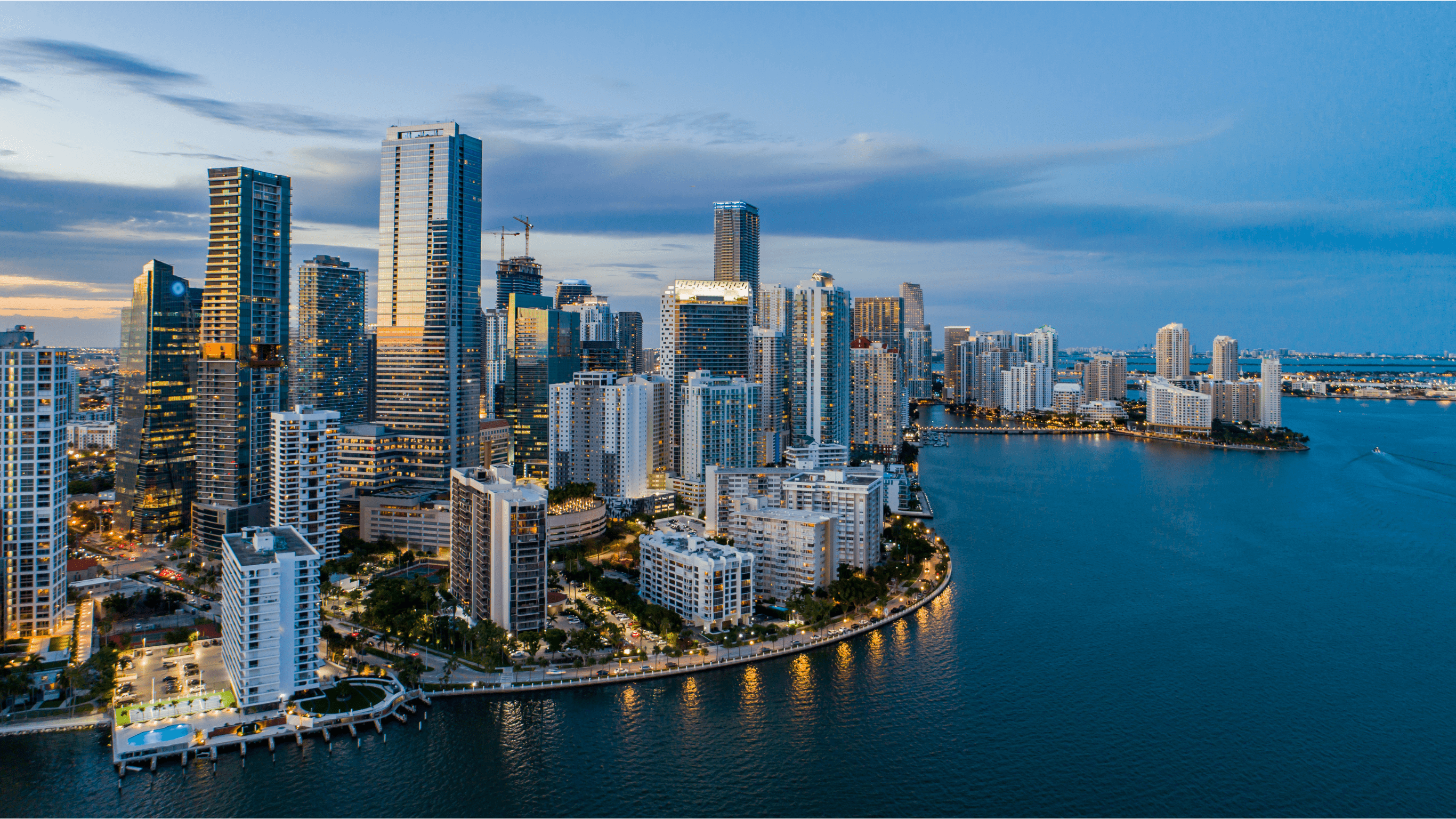 Aerial view of a modern city skyline with high-rise buildings along the waterfront at dusk, reflecting lights on the calm water.